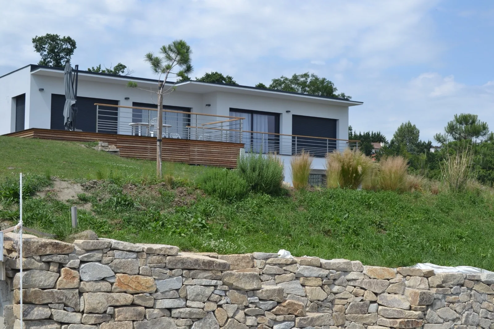 Maison neuve à toit plat, terrasse en bois et garde-corps, sur un terrain en pente au-dessus d'un mur en pierre sèche